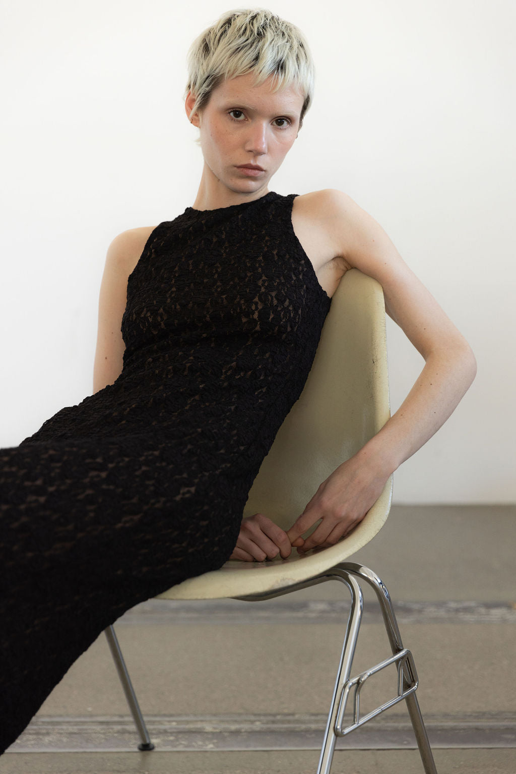 A person with short blonde hair wears a sleeveless black lace maxi dress while sitting on a light-colored chair.