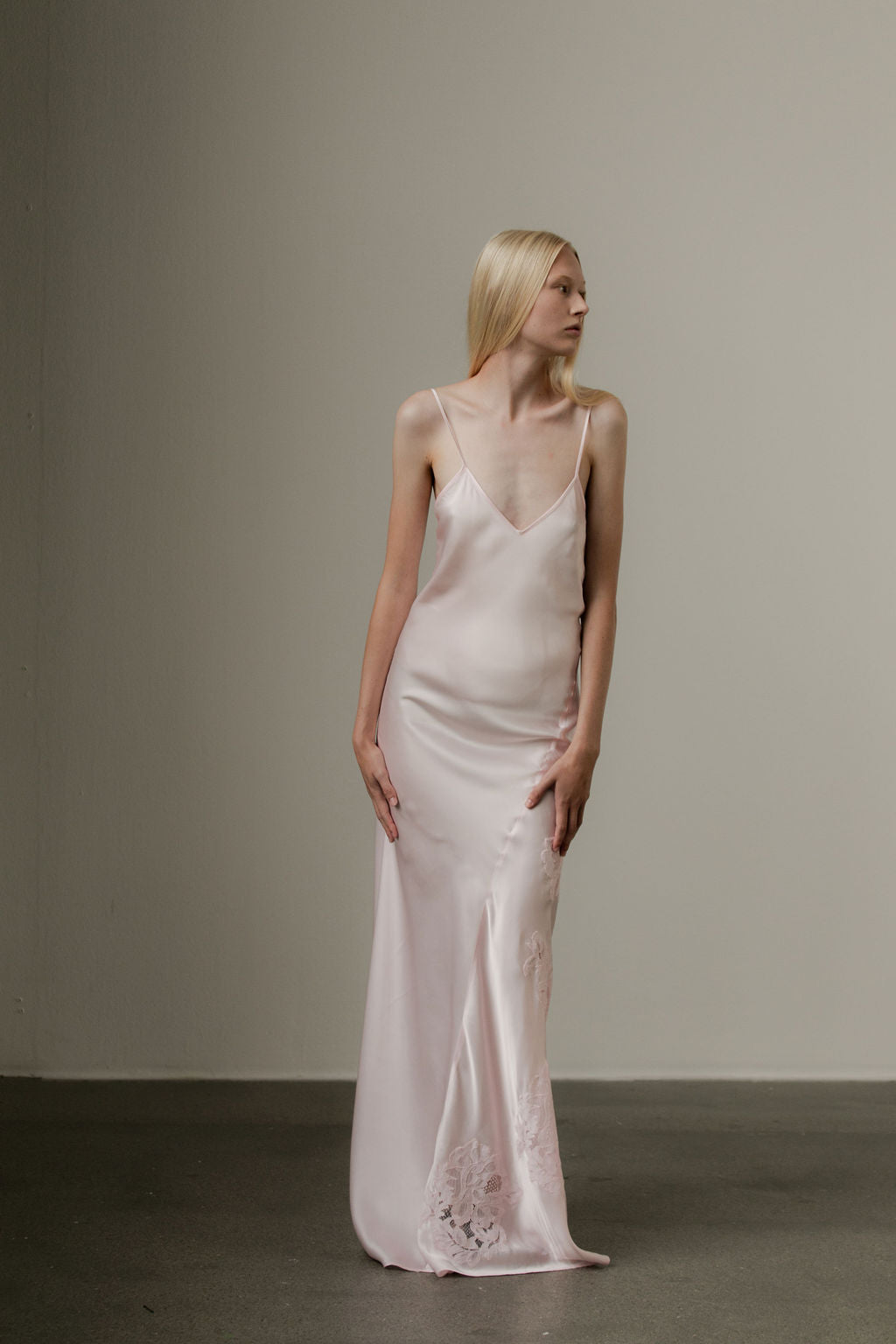 A model wearing a pale pink, floor-length silk slip dress with lace detailing on the lower side.