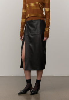 Wearing a brown and mustard striped sweater, the person pairs it with the Herskind ApS Elizabeth Leather Skirt—a black lamb leather midi skirt featuring a front slit—along with black sheer socks and black loafers, while standing against a plain background.