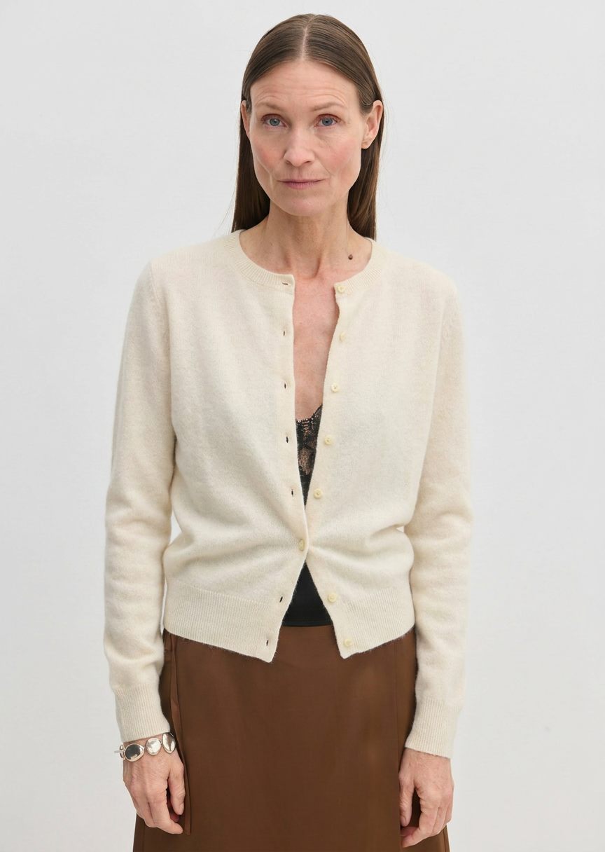A woman wears a cream-colored, long-sleeved cashmere crew-neck Herskind ApS Roselle Knit Cardigan buttoned at the front over a black camisole.