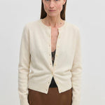 A woman wears a cream-colored, long-sleeved cashmere crew-neck Herskind ApS Roselle Knit Cardigan buttoned at the front over a black camisole.