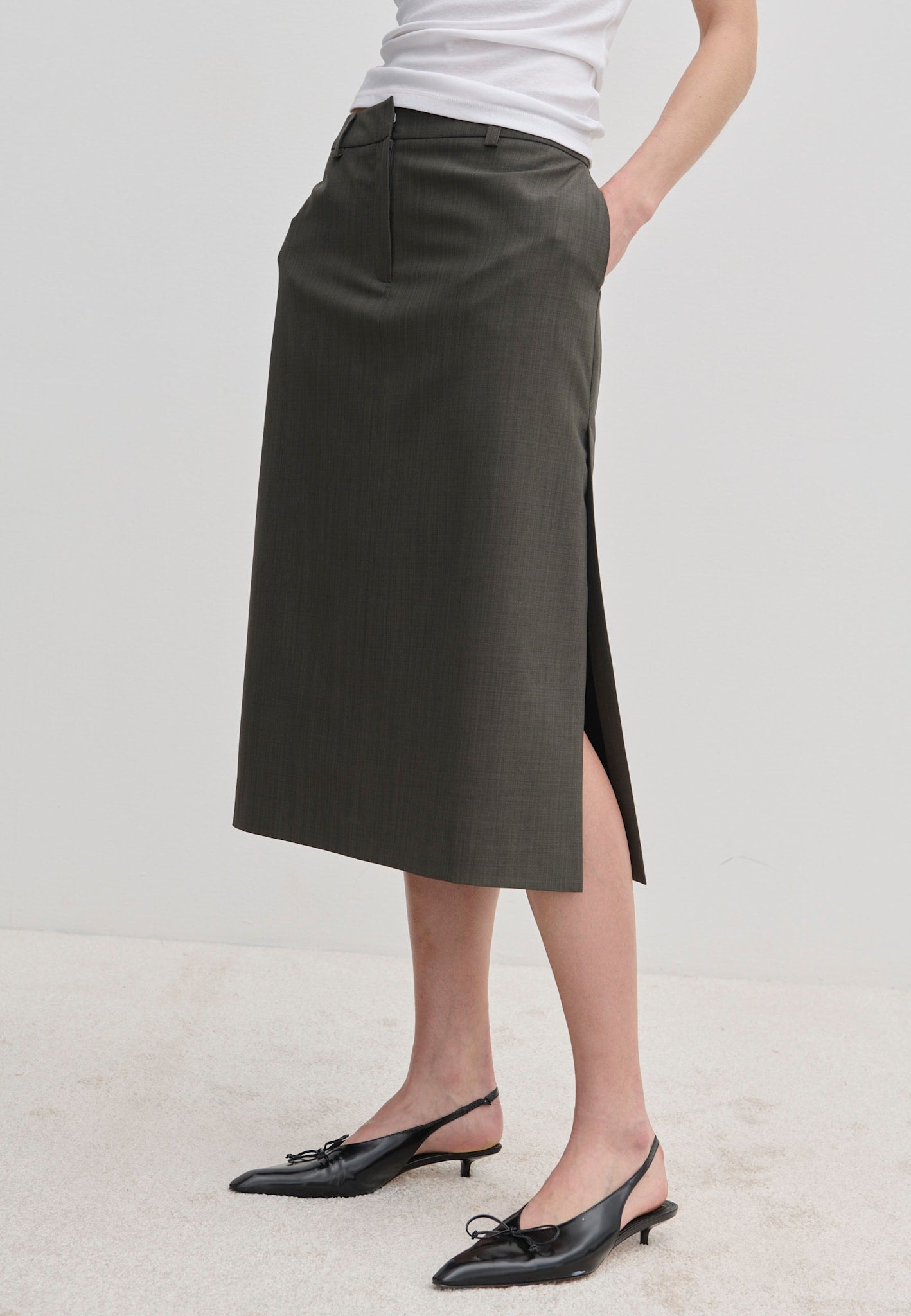 A person wearing a dark grey, straight-silhouette Herskind ApS Collin Mid waist Skirt with a side slit and black kitten-heel slingback shoes.