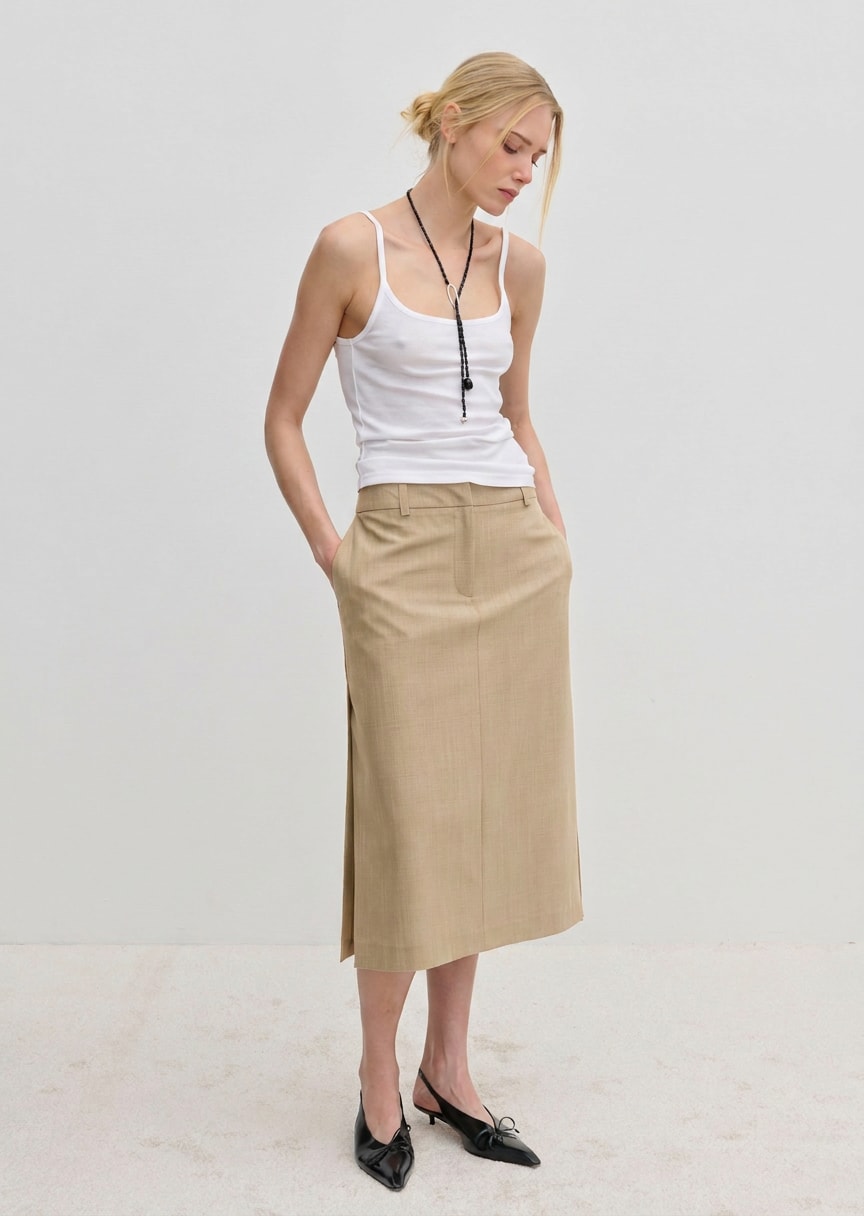 A woman wears a white tank top and a tan, straight silhouette Collin Mid waist Skirt by Herskind ApS, featuring a midi length and a side vent.