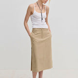 A woman wears a white tank top and a tan, straight silhouette Collin Mid waist Skirt by Herskind ApS, featuring a midi length and a side vent.