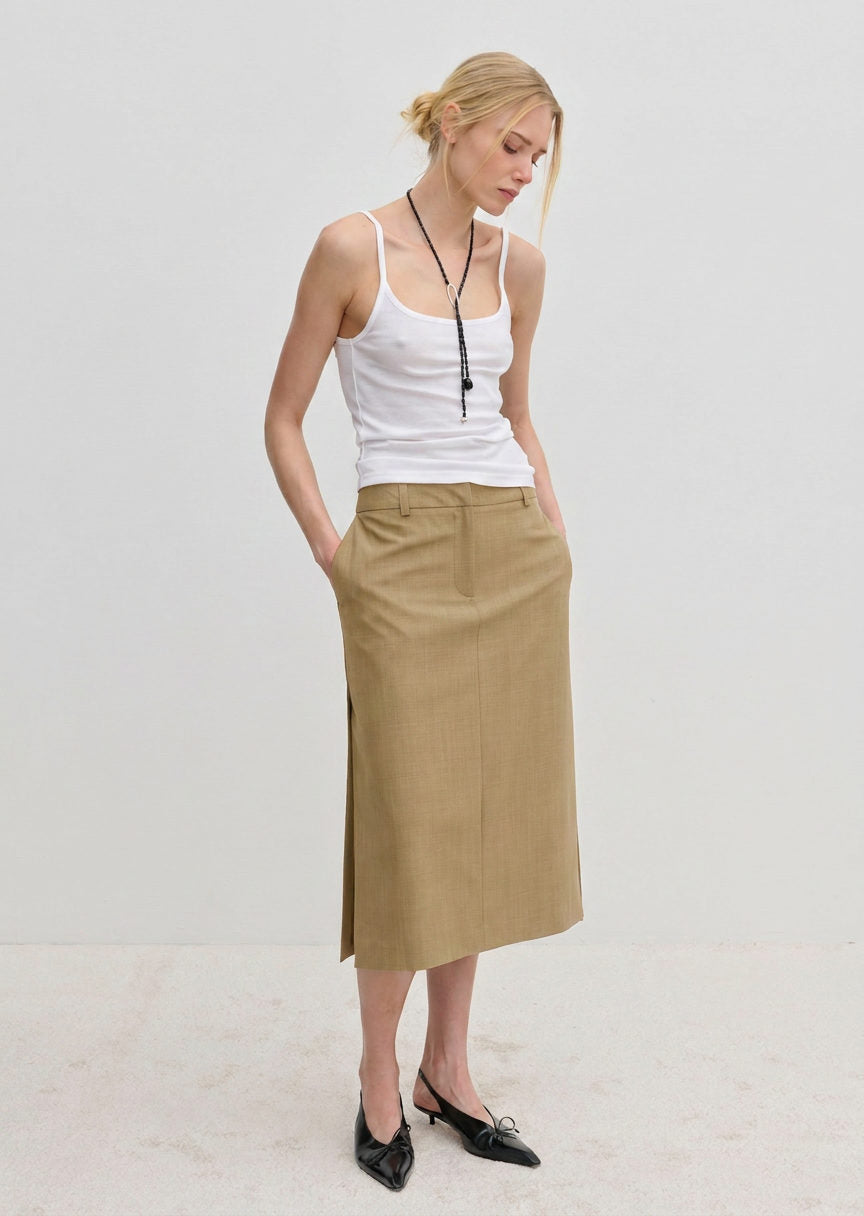 A person wearing a white spaghetti strap tank top and a tan, midi-length Herskind ApS Collin Mid waist Skirt with a side slit.