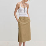 A person wearing a white spaghetti strap tank top and a tan, midi-length Herskind ApS Collin Mid waist Skirt with a side slit.