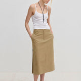 A person wearing a white spaghetti strap tank top and a tan, midi-length Herskind ApS Collin Mid waist Skirt with a side slit.