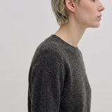 A person in profile wearing an Elena Knit Blouse by Herskind ApS, a dark grey long-sleeved crew-neck knit blouse with a ribbed hem.