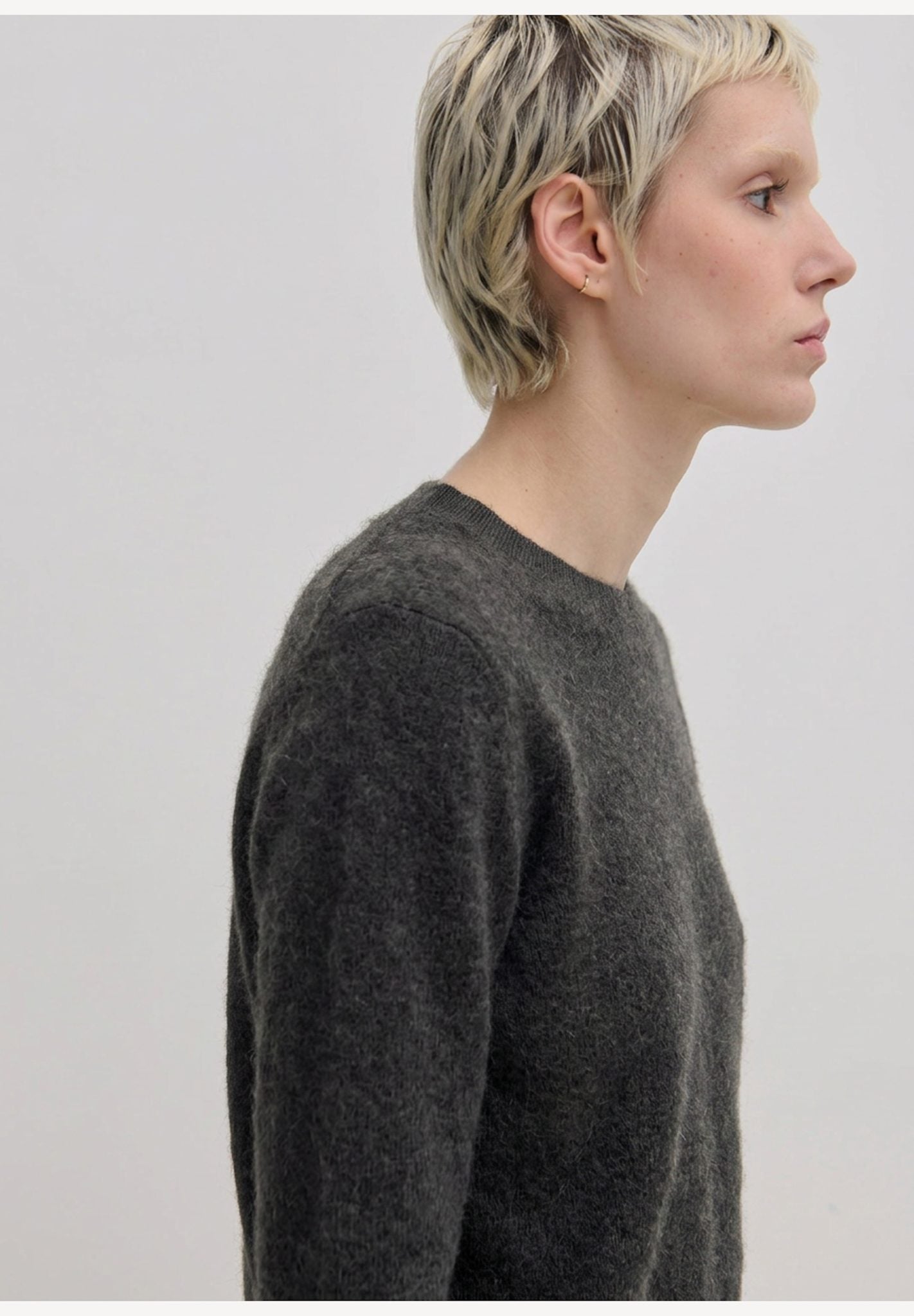 A person in profile wearing an Elena Knit Blouse by Herskind ApS, a dark grey long-sleeved crew-neck knit blouse with a ribbed hem.