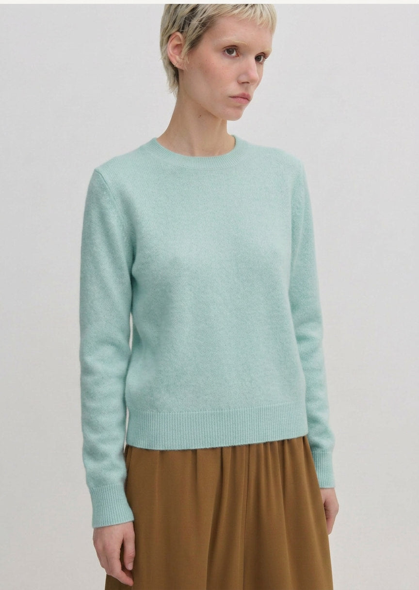 A person wearing a pale green Herskind ApS Elena Knit Blouse with a crew neck and a ribbed hem.