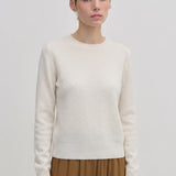 A person wearing an Elena Knit Blouse by Herskind ApS, featuring a long-sleeved, cream-colored crew-neck design with a ribbed hem.