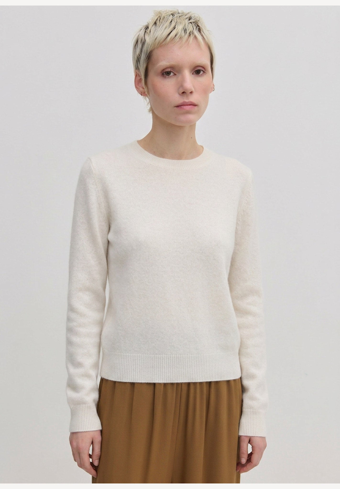 A person wearing an Elena Knit Blouse by Herskind ApS, featuring a long-sleeved, cream-colored crew-neck design with a ribbed hem.