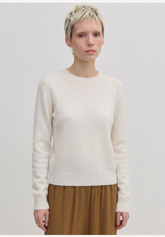 A person wearing an Elena Knit Blouse by Herskind ApS, featuring a long-sleeved, cream-colored crew-neck design with a ribbed hem.