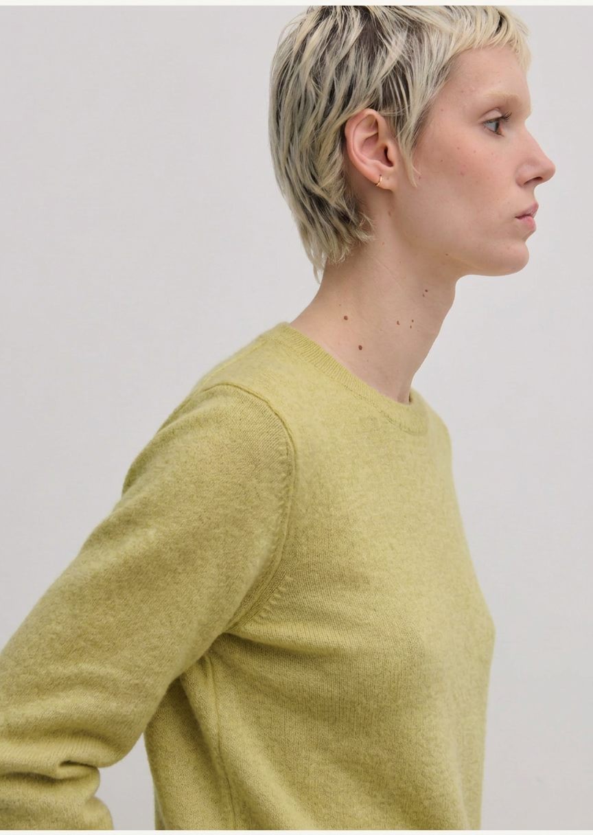 A person with short blonde hair wears a lime green Herskind ApS Elena Knit Blouse, a long-sleeved, crew-neck knit top with a ribbed hem.
