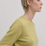 A person with short blonde hair wears a lime green Herskind ApS Elena Knit Blouse, a long-sleeved, crew-neck knit top with a ribbed hem.