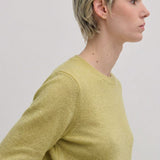 A person with short blonde hair wears a lime green Herskind ApS Elena Knit Blouse, a long-sleeved, crew-neck knit top with a ribbed hem.