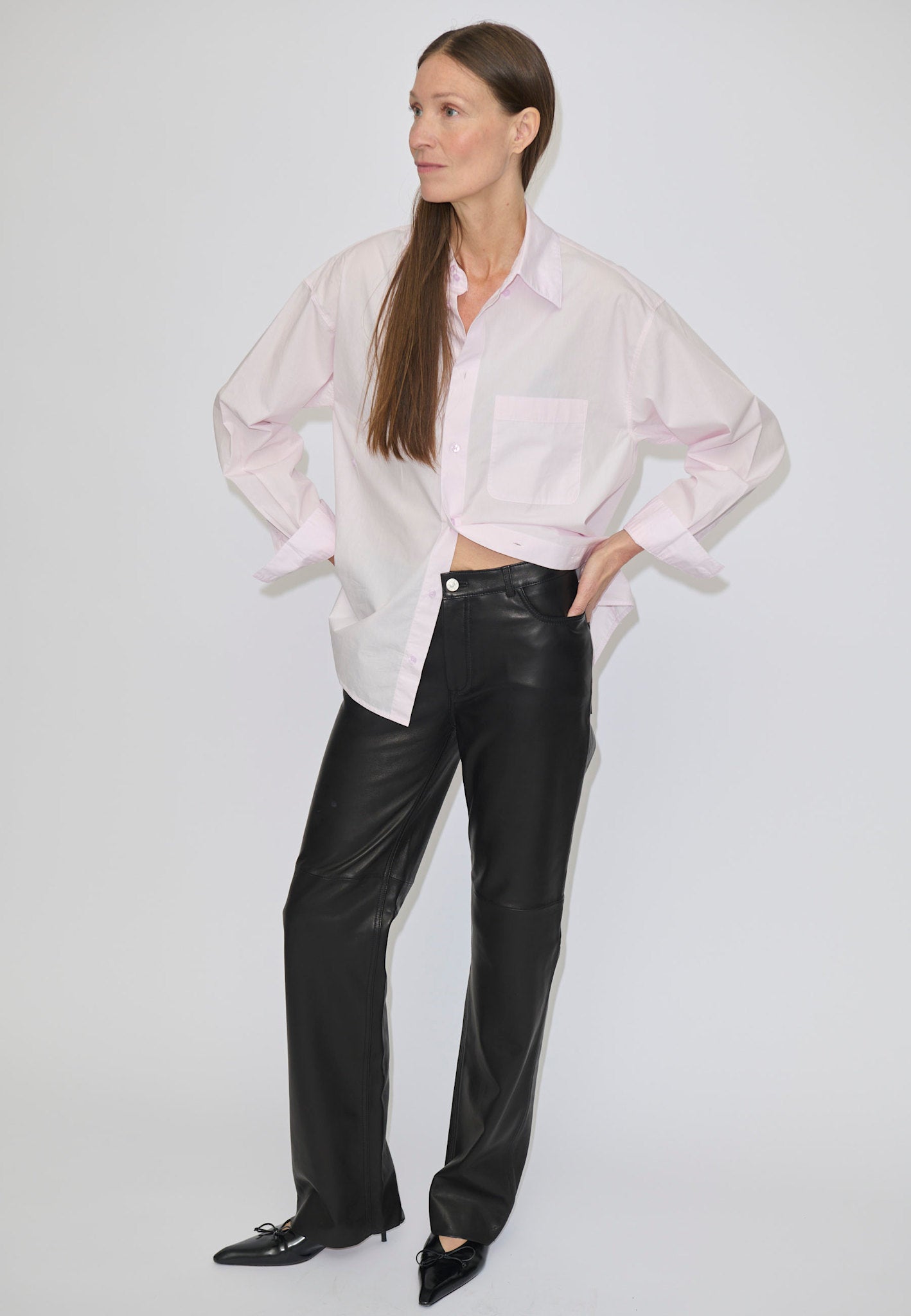 A person wearing a pale pink button-down shirt and high-waisted, straight-fit black Cooper Leather Jeans by Herskind ApS, with their hands on their hips.