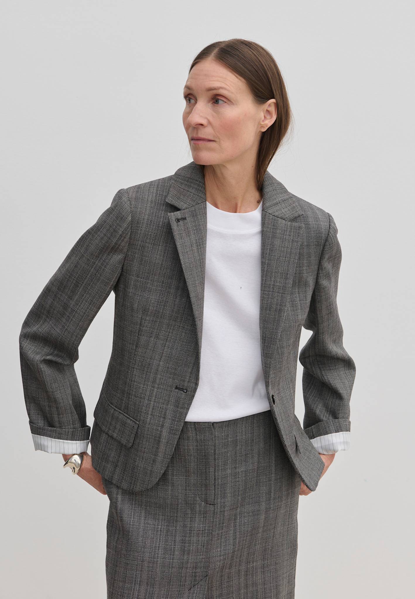 A person wearing a grey plaid Dakota Fitted Blazer by Herskind ApS over a white t-shirt, with the sleeves rolled up to reveal white striped lining.