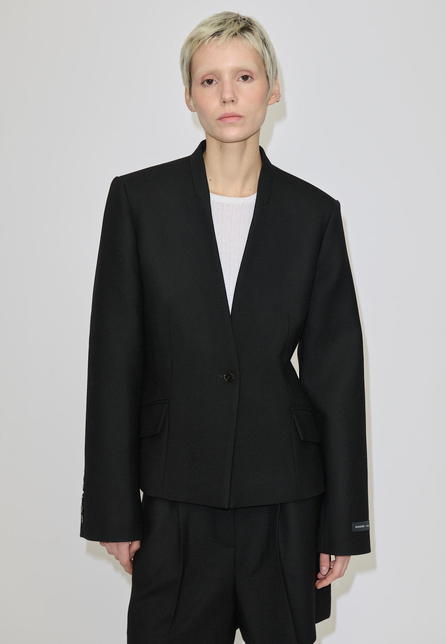 A person wears a black, single-button, fitted Higgins Fitted Blazer by Herskind ApS with a notched neckline over a white top.