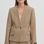A person wearing the tan, collarless Higgins Fitted Blazer by Herskind ApS, crafted from recycled polyester with a single-button closure and two flap pockets.