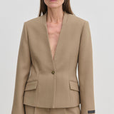A person wearing the tan, collarless Higgins Fitted Blazer by Herskind ApS, crafted from recycled polyester with a single-button closure and two flap pockets.