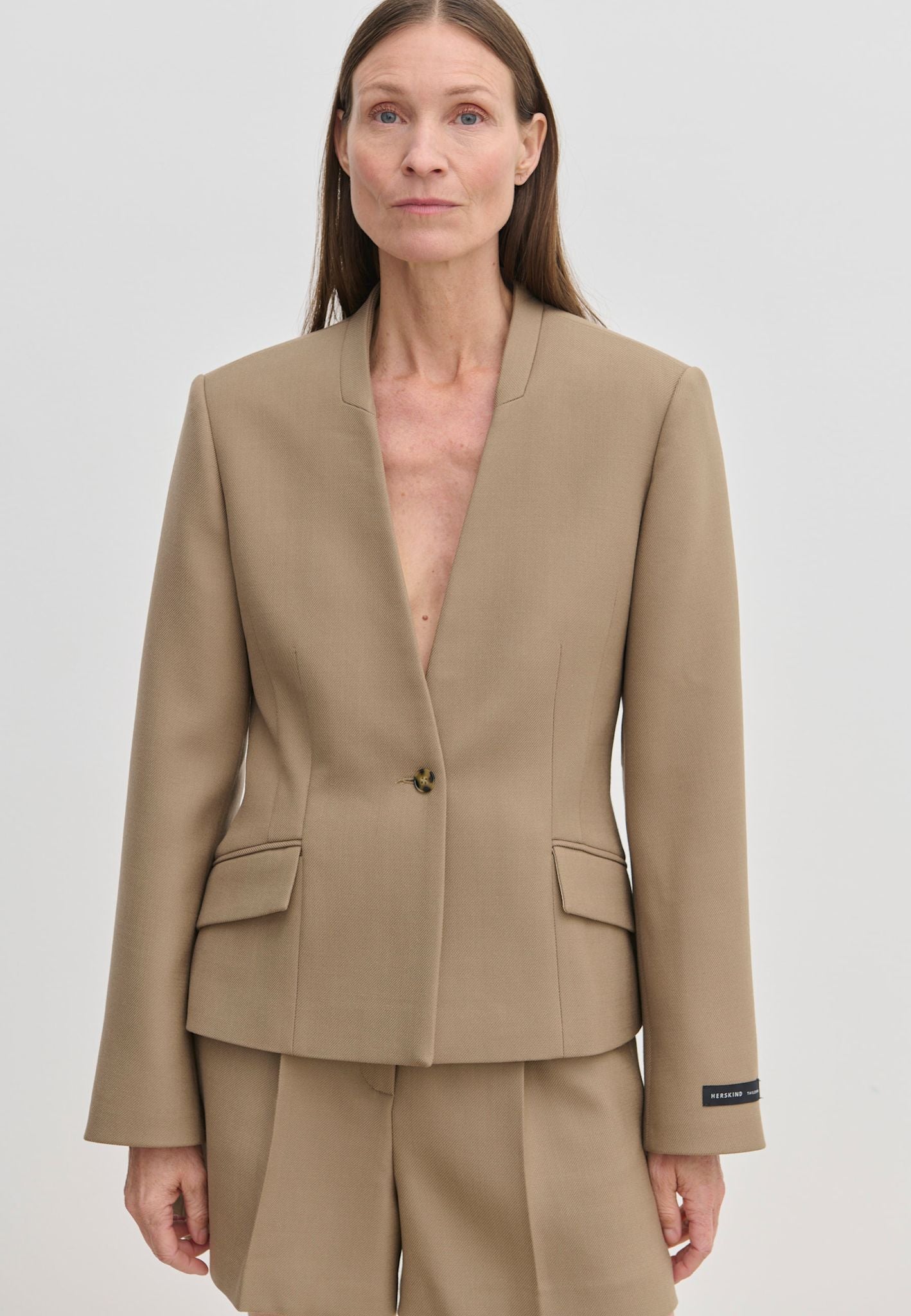 A person wearing the tan, collarless Higgins Fitted Blazer by Herskind ApS, crafted from recycled polyester with a single-button closure and two flap pockets.