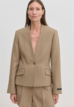 A person wearing the tan, collarless Higgins Fitted Blazer by Herskind ApS, crafted from recycled polyester with a single-button closure and two flap pockets.