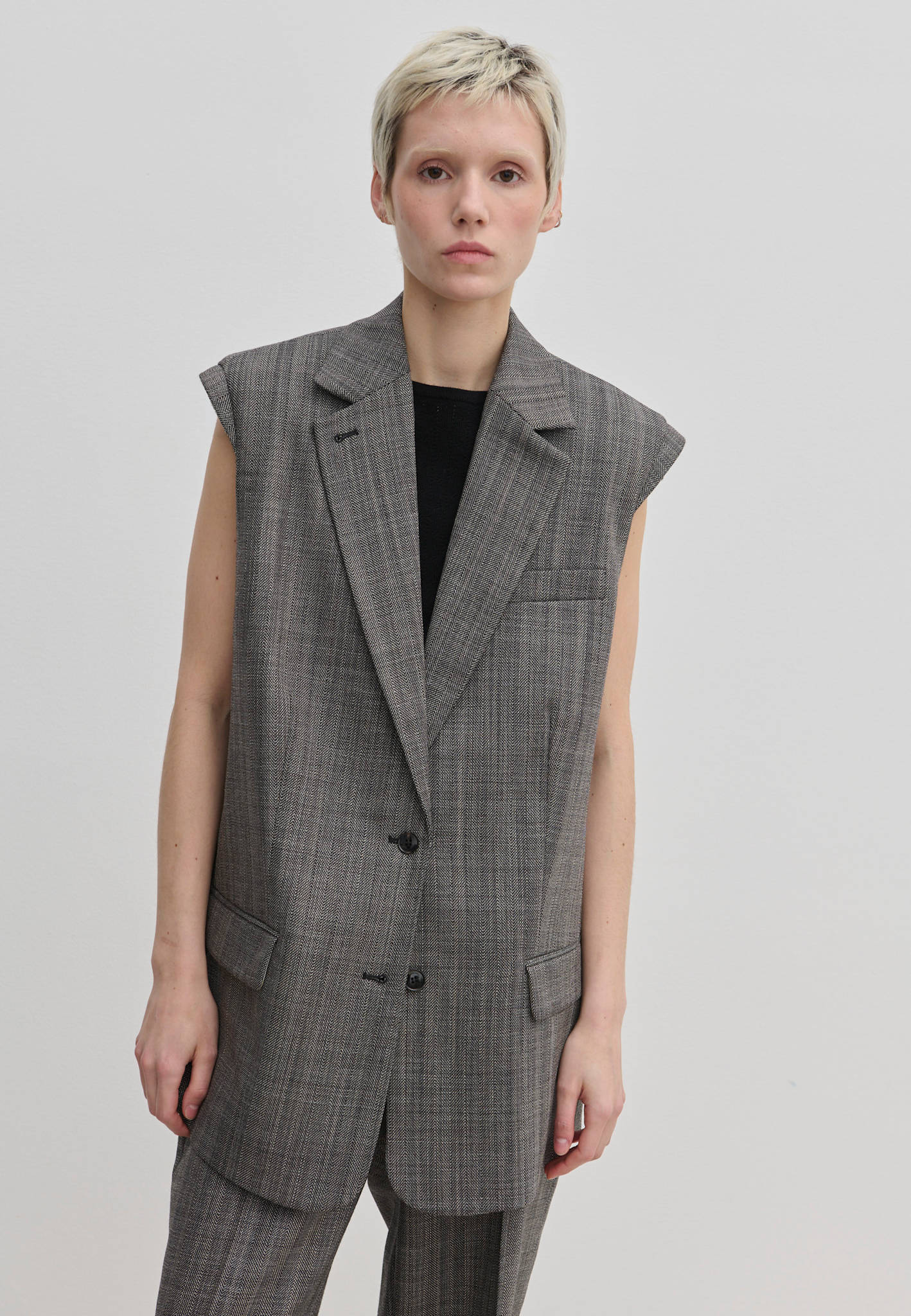 A person wearing a structured, sleeveless grey plaid Herskind ApS Cohen Sleeveless Blazer over a black top.