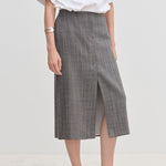 A person wearing a grey plaid Duncan Mid waist Skirt by Herskind ApS with a front slit, a white t-shirt, and pale shoes.