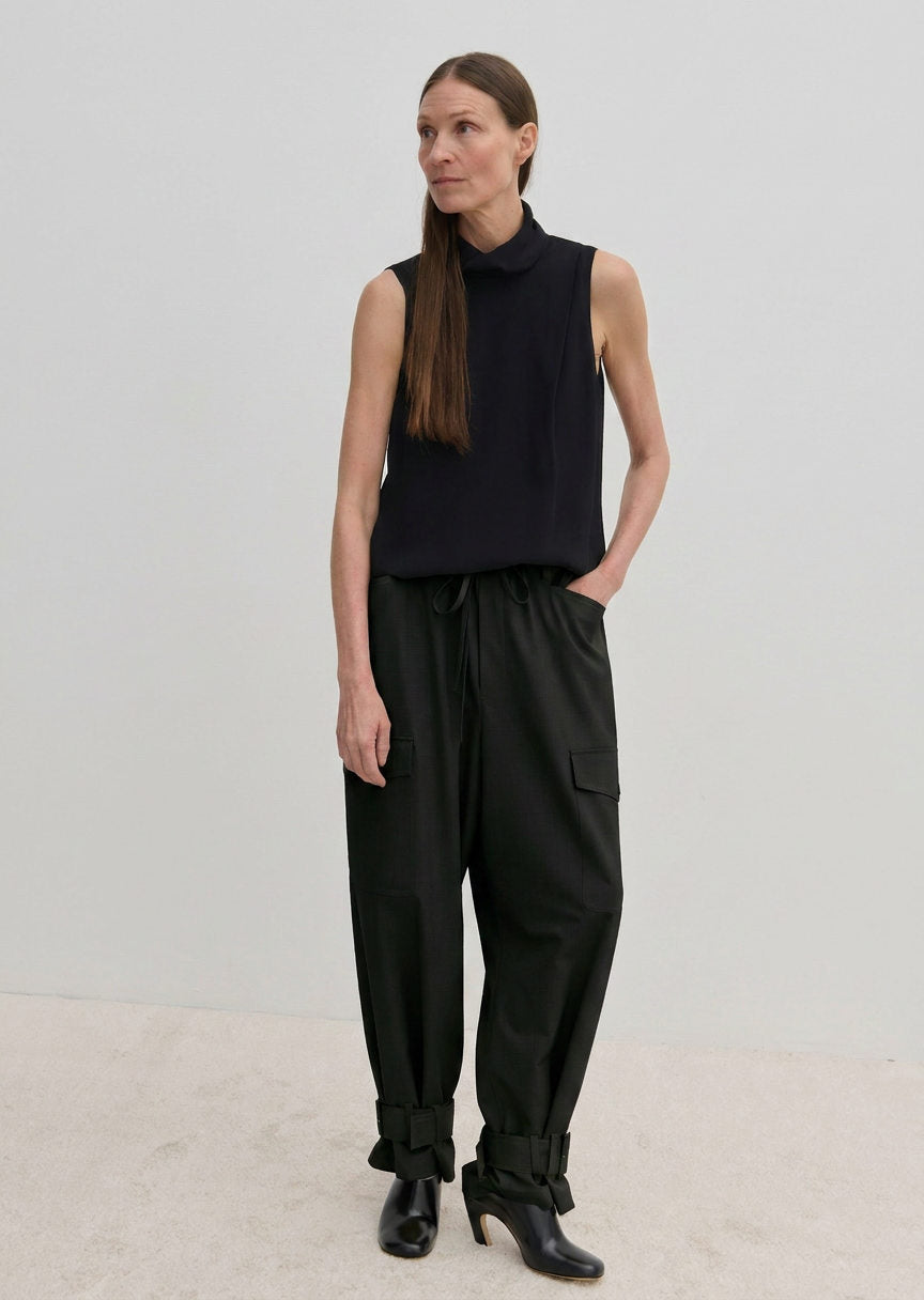 A person wears a sleeveless black top and Herskind ApS Maddie Mid waist Baggy Pants, which feature a relaxed-fit, baggy silhouette with side pockets and buckled ankle cuffs.