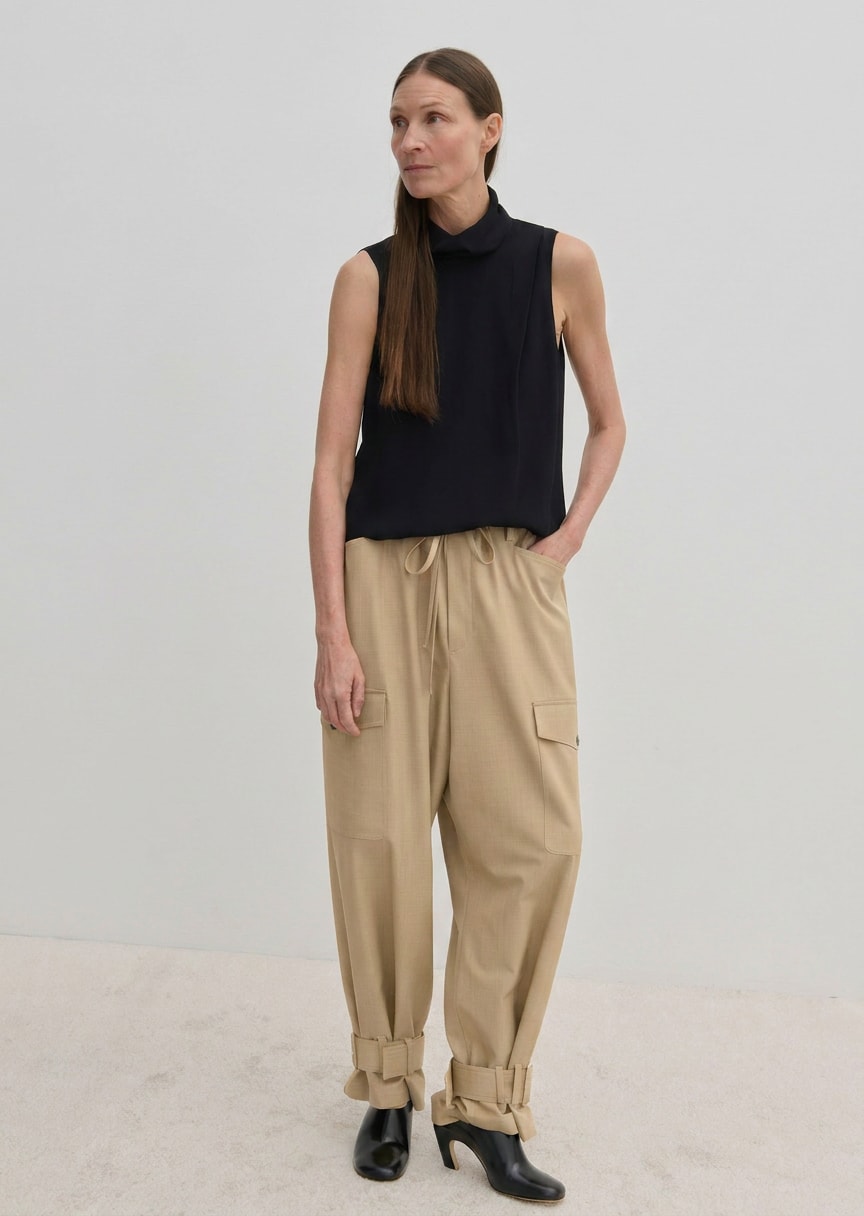 A woman wears a sleeveless black mock-neck top and beige Herskind ApS Maddie Mid waist Baggy Pants with buckled hems.