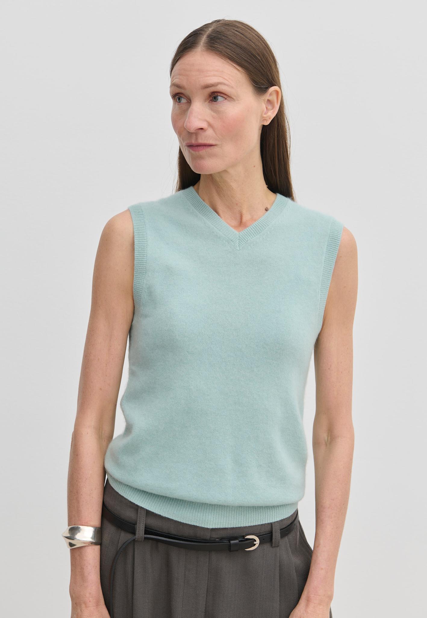 A woman wears a light blue V-neck Fabien Knit Vest by Herskind ApS over dark trousers.