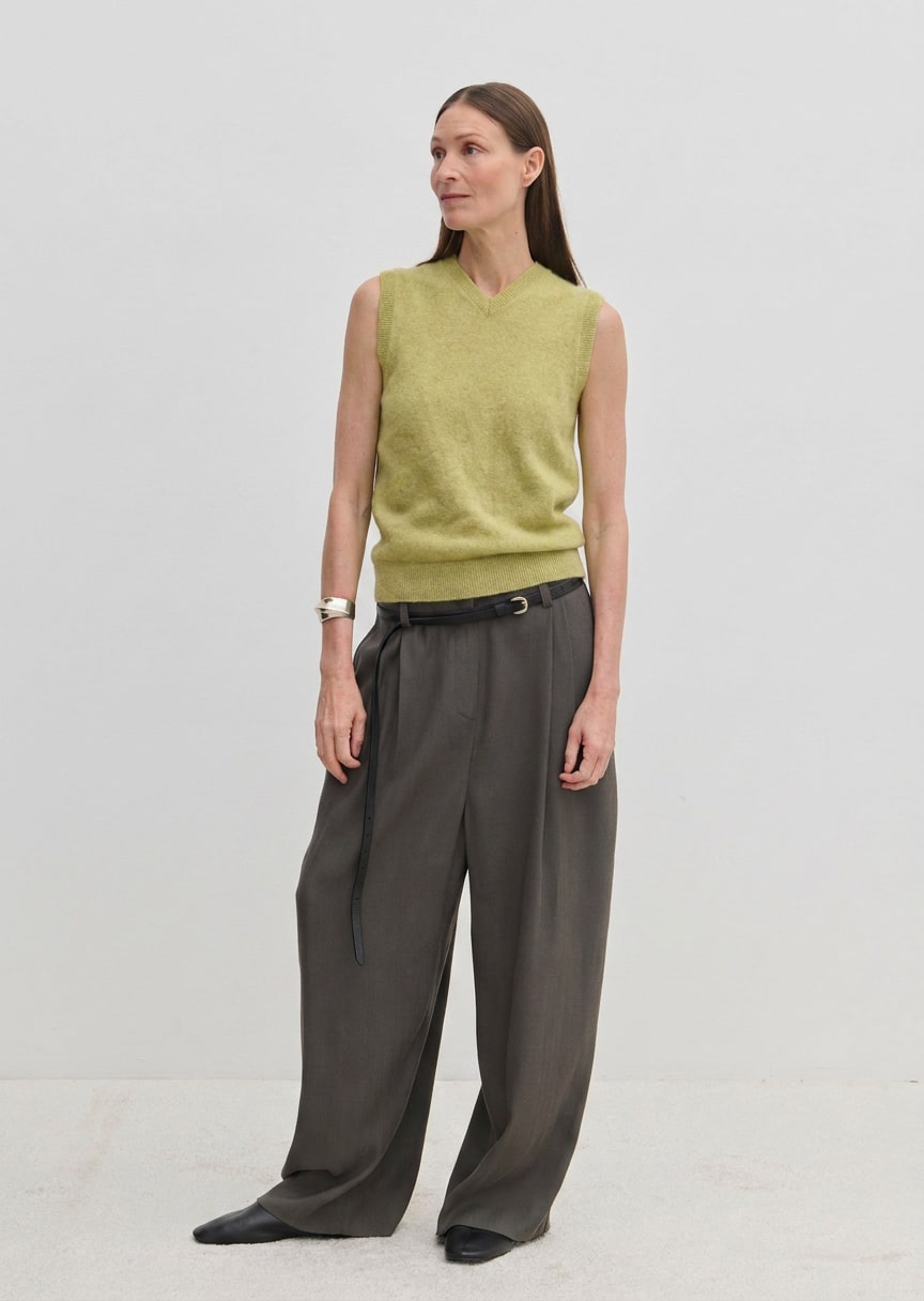 A person wearing a pale green Fabien Knit Vest tucked into charcoal wide-leg trousers with a black belt.