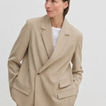 A person wearing an Amalfi Regular Blazer by Herskind ApS, a beige double-breasted style with structured shoulders and flap pockets over a white crew-neck shirt.