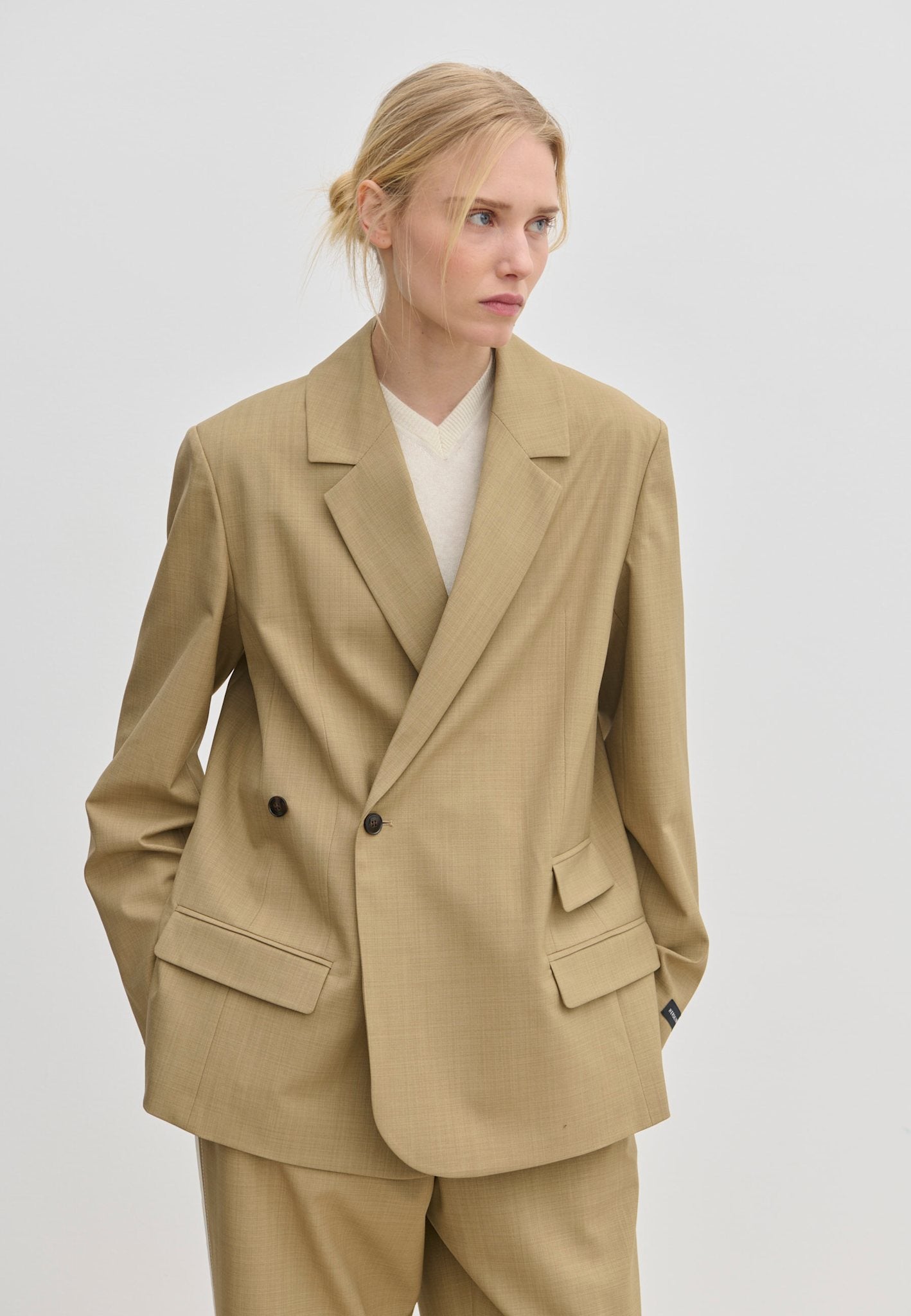A person wearing a tan Amalfi Regular Blazer by Herskind ApS with structured shoulders over a white V-neck sweater.