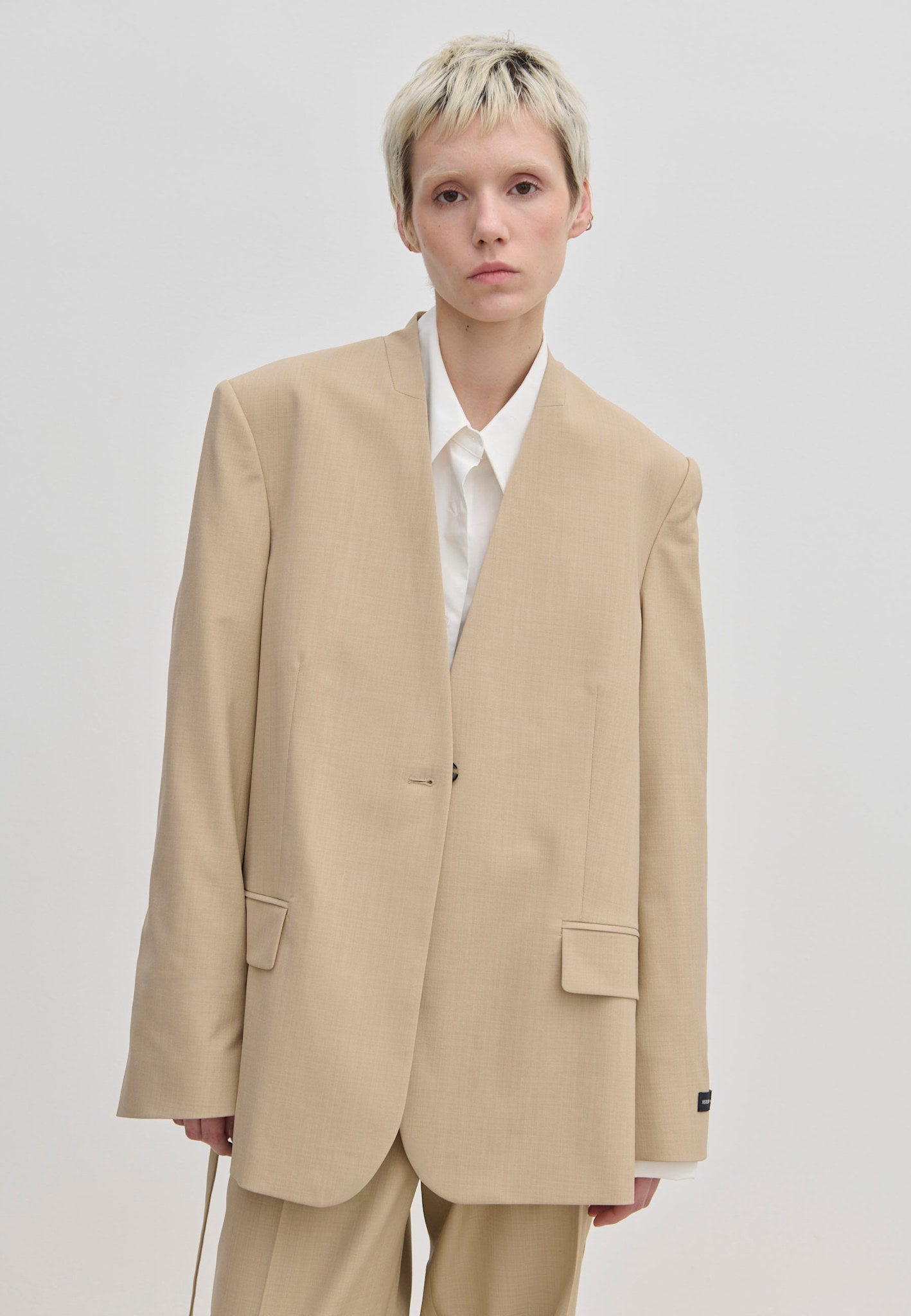 A model wearing an oversized, modern beige Herskind ApS Clarissa Regular Blazer with structured shoulders over a white collared shirt.