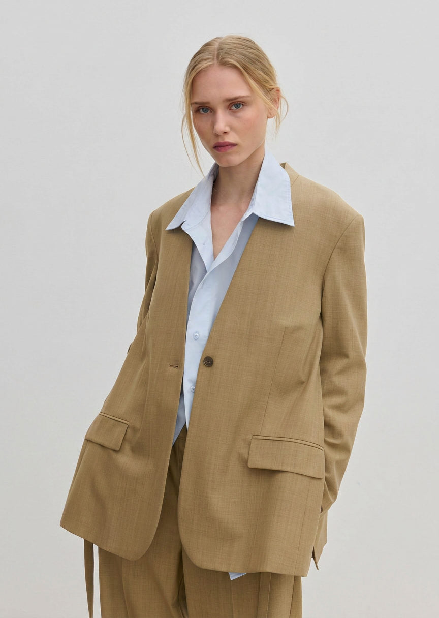 A person wears a modern, tan, single-button Clarissa Regular Blazer by Herskind ApS over a light blue collared shirt.