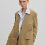 A person wears a modern, tan, single-button Clarissa Regular Blazer by Herskind ApS over a light blue collared shirt.