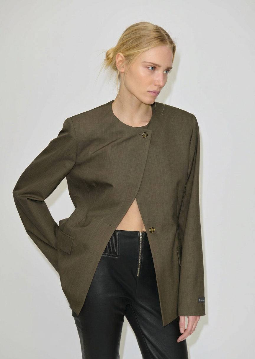 A person wears an olive green, collarless Robyn Fitted Blazer by Herskind ApS, featuring two buttons and styled open over black trousers with a front zipper.
