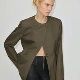 A person wears an olive green, collarless Robyn Fitted Blazer by Herskind ApS, featuring two buttons and styled open over black trousers with a front zipper.