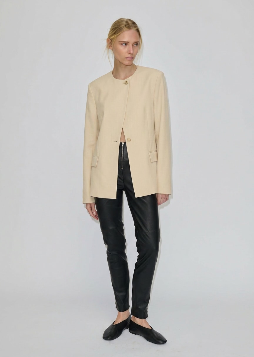 A model wearing a beige collarless Robyn Fitted Blazer by Herskind ApS with structured shoulders, black fitted trousers, and black pointed-toe flats.