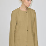 A person wearing a tan, collarless Robyn Fitted Blazer by Herskind ApS, featuring structured shoulders, two visible front buttons, and two flap pockets.