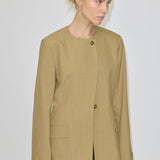 A person wearing a tan, collarless Robyn Fitted Blazer by Herskind ApS, featuring structured shoulders, two visible front buttons, and two flap pockets.