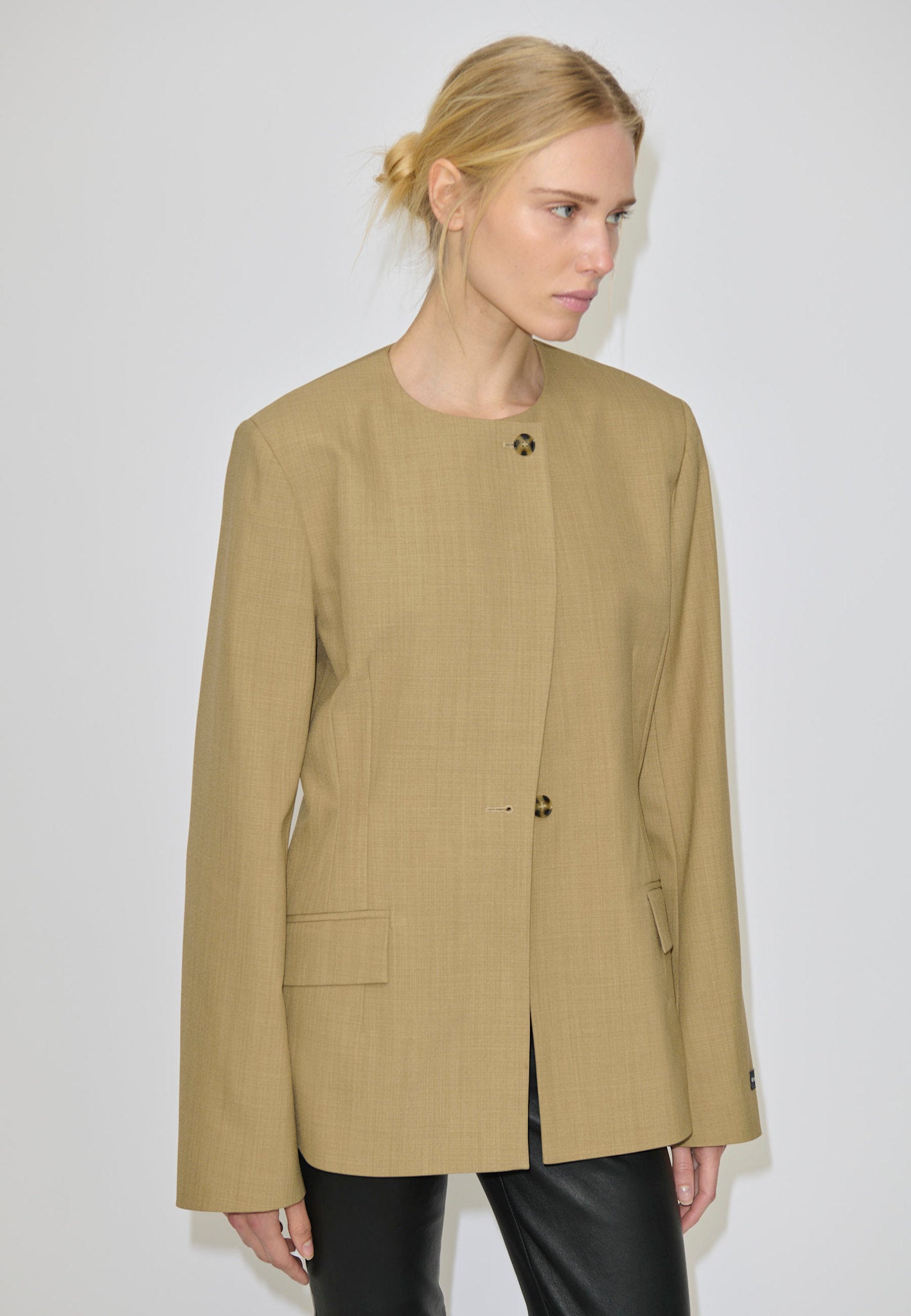 A person wearing a tan, collarless Robyn Fitted Blazer by Herskind ApS, featuring structured shoulders, two visible front buttons, and two flap pockets.