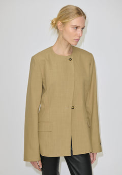 A person wearing a tan, collarless Robyn Fitted Blazer by Herskind ApS, featuring structured shoulders, two visible front buttons, and two flap pockets.