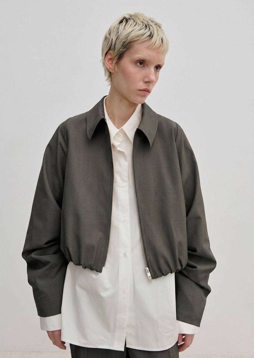 A person wearing a charcoal gray, cropped-length Stella Jacket by Herskind ApS over a white button-down shirt.