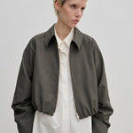 A person wearing a charcoal gray, cropped-length Stella Jacket by Herskind ApS over a white button-down shirt.