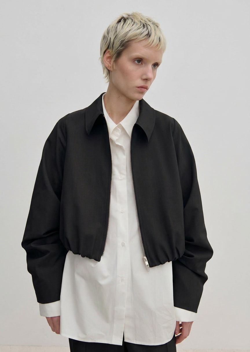 A person wearing a relaxed fit, cropped black Stella Jacket by Herskind ApS over a long-sleeved white button-down shirt.