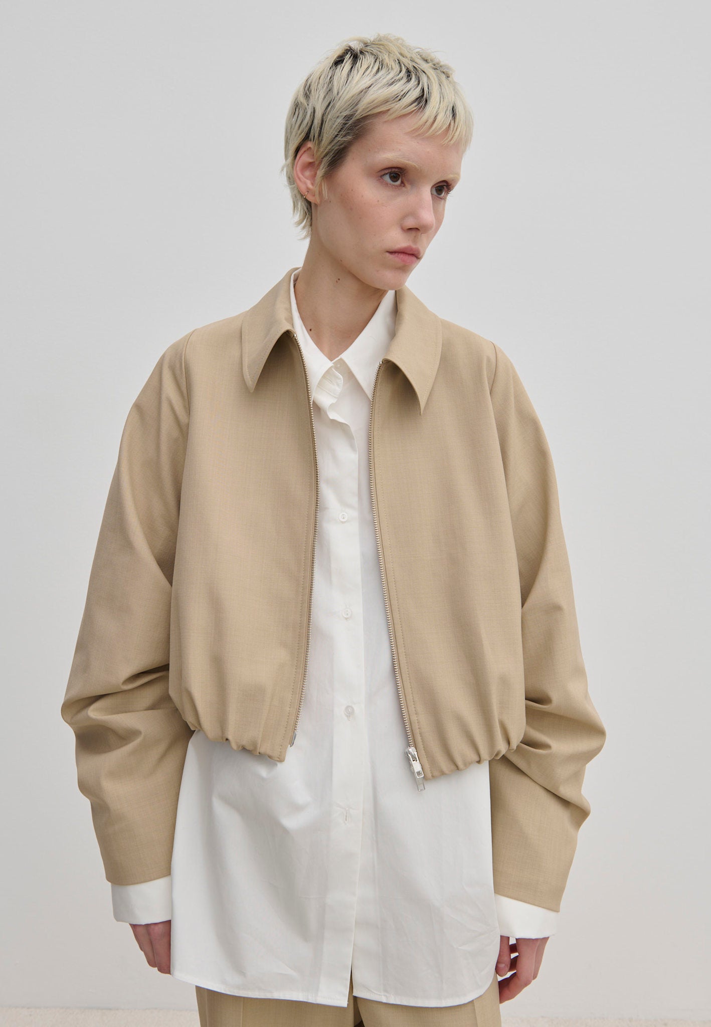 A person wearing a relaxed fit, cropped-length beige Stella Jacket by Herskind ApS with a wide collar over a white button-down shirt.