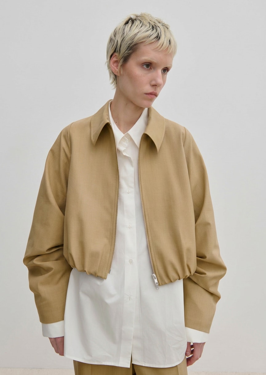 A person wearing a relaxed fit, cropped tan Stella Jacket by Herskind ApS over a long-sleeved white button-down shirt.